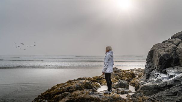 Starsza kobieta spogląda w gęstą mgłę nad Oceanem Spokojnym w zatoce Cox w Parku Narodowym Pacific Rim na wyspie Vancouver, Kolumbia Brytyjska, Kanada - Zdjęcie, obraz