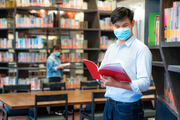 Dwóch studentów azjatyckich uniwersytetów noszących maski twarzy i stojących w bibliotece społecznej odległości od innych 6 stóp, aby uniknąć rozprzestrzeniania się koronawirusu na Uniwersytecie. - Zdjęcie, obraz