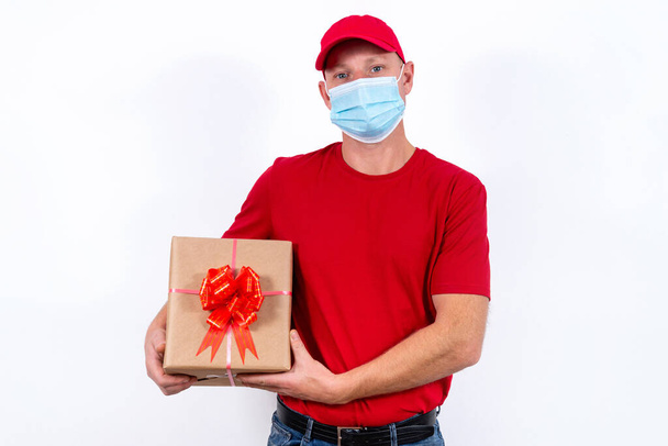 Safe delivery of gifts for holidays. A courier in red uniform and protective medical mask holds box with a bow. Contactless remote gift orders in quarantine during the coronavirus pandemic. - Photo, image