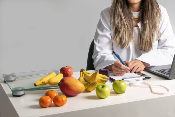 Γραφείο γυναίκας διατροφολόγου στο γραφείο της, γράφει πρόγραμμα διατροφής που δείχνει υγιεινά λαχανικά και φρούτα. Υγιεινή και διατροφή. Τρόπος ζωής. - Φωτογραφία, εικόνα