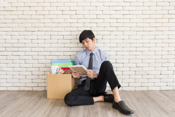 Aasialainen mies istui valitettavasti puulattialla toimistossa potkujen jälkeen, säilyttäen henkilökohtaisia tavaroita pahvilaatikoissa.. - Valokuva, kuva