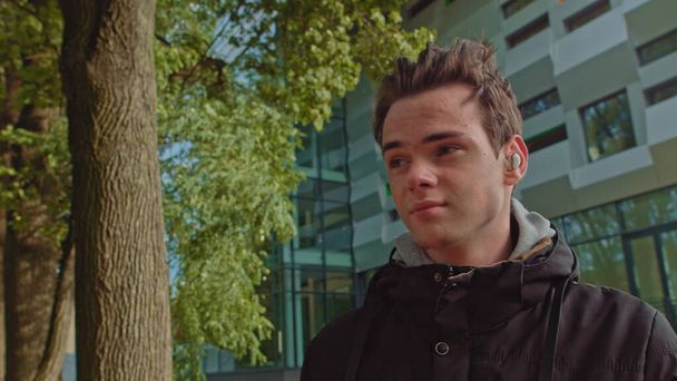 Mladý muž mluví do telefonu pomocí bezdrátových sluchátek venku, když jde po ulici. Mladíku, teenager nosí moderní bezdrátová sluchátka. Portrét mladého muže. - Fotografie, Obrázek