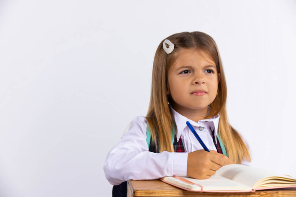  Little girl in a school uniform sitting at a table on a white background with blank space. The schoolgirl is doing her homework and looking away thoughtfully. Education concept. High quality photo - Valokuva, kuva