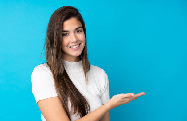 tiener meisje over geïsoleerde blauwe achtergrond presenteren van een idee terwijl het kijken glimlachend naar - Foto, afbeelding