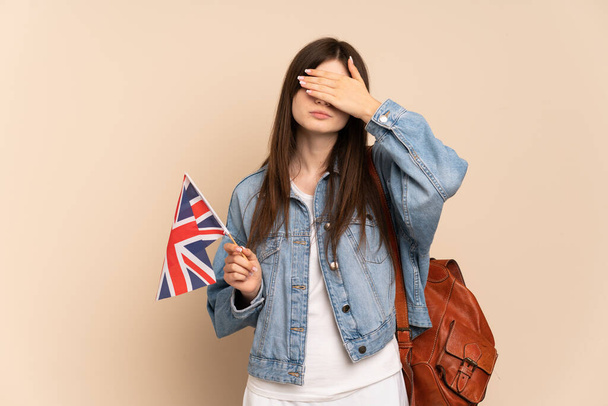 Молода українська дівчина, що тримає прапор Великої Британії, ізольована на бежевому фоні, закриває очі руками. Не хочу нічого бачити. - Фото, зображення