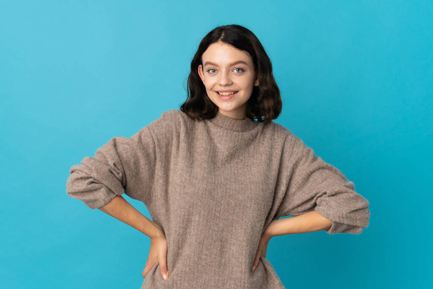 Teenager Ukrainian girl isolated on blue background posing with arms at hip and smiling - Photo, Image