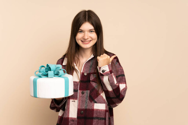 Young Ukrainian girl holding a big cake isolated on beige background pointing to the side to present a product - Foto, Bild