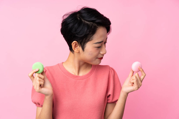 Menina asiática sobre isolado fundo rosa segurando macarons franceses coloridos e olhando-o - Foto, Imagem