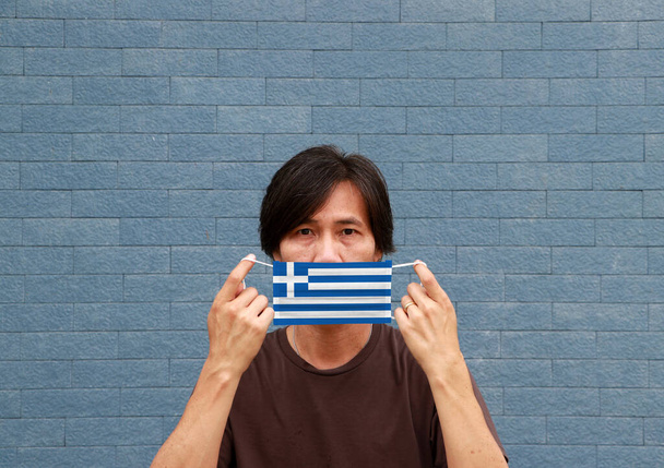 彼の手の中にギリシャの旗のパターンを持つ男と衛生的なマスクと青い壁の背景に彼の顔をカバーするためにそれを上げる。マスクは小さな粒子やウイルスコロナから非常に良い保護です。. - 写真・画像
