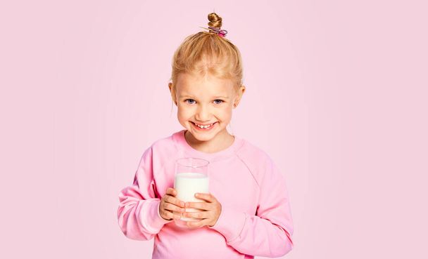 Elinde büyük bir bardak süt tutan tatlı küçük kız. 4-5 yaşındaki çocuk stüdyoda pembe arka planda poz veriyor. - Fotoğraf, Görsel
