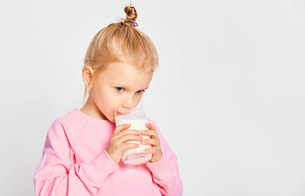 Słodka dziewczynka pijąca dużą szklankę mleka. Kid 4-5 lat pozowanie w studio na szarym tle - Zdjęcie, obraz