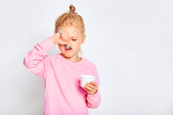 Schattig klein meisje dat lekkere yoghurt eet op een grijze achtergrond. 4-5 jaar oud kind poseren in de studio in een roze sweater. Ruimte voor tekst - Foto, afbeelding