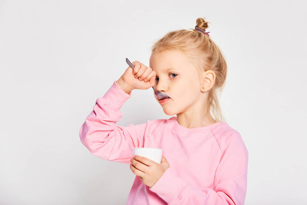 Nettes kleines Mädchen, das leckeren Joghurt auf grauem Hintergrund isst. 4-5 jährige Kinder posieren im Studio in einem rosafarbenen Sweatshirt. Raum für Text - Foto, Bild