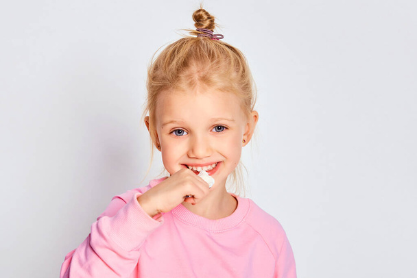 Cute little girl eating tasty yogurt on gray background. 4-5 year old kid posing in studio in a pink sweatshirt. Space for text - Foto, Imagem
