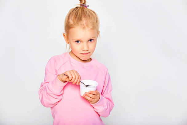 Linda niña comiendo sabroso yogur sobre fondo gris. Niño de 4-5 años posando en un estudio con una sudadera rosa. Espacio para texto - Foto, imagen
