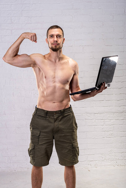 A man with a naked torso holds a laptop and demonstrates biceps. Remote training. Trainer. - Fotoğraf, Görsel