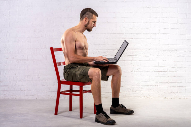 A man with a naked torso sits on a chair and holds a laptop. Remote training. Trainer. - Photo, Image