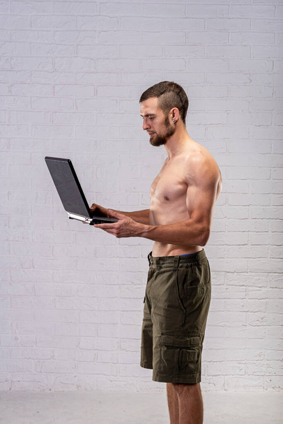 A man with a naked torso holds a laptop. Remote training. Trainer. - Zdjęcie, obraz