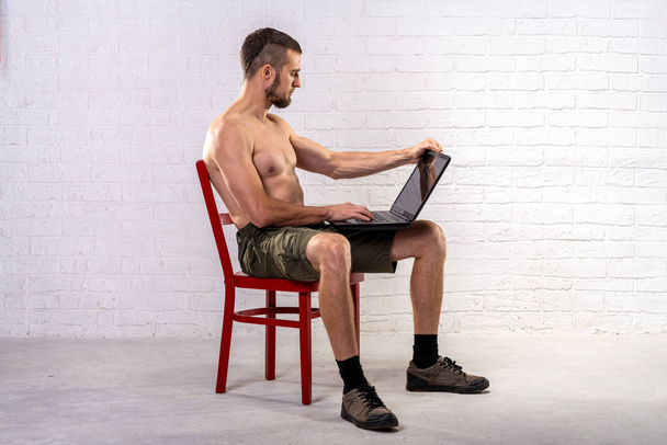 A man with a naked torso sits on a chair and holds a laptop. Remote training. Trainer. - Фото, изображение