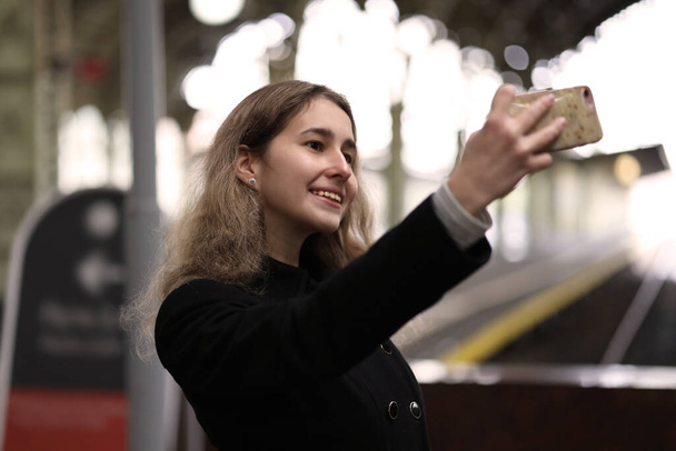 young happy girl makes selfie on a mobile phone at the train station on the platform - Foto, imagen
