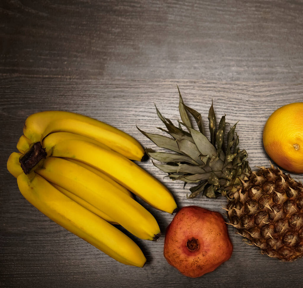  one pineapple, grapefruit, pomegranate and fresh bunch of bananas. top view. black wooden table background.empty copy space. row healthy food - Photo, image