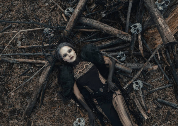 Jovem mulher na imagem de bruxa encontra-se no chão em vestido preto e coroa na cabeça perto de árvores caídas na floresta e crânios humanos. Vista superior. - Foto, Imagem