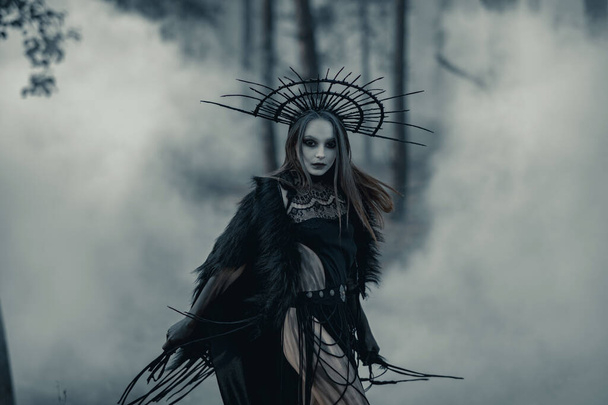 Young woman in image of witch stands in black dress and crown on her head in dark and foggy forest. - Foto, Imagen