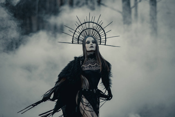 Young woman in image of witch stands in black dress and crown on her head in dark and foggy forest. - Фото, зображення