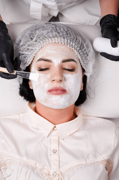 Κοντινό πλάνο της cosmetologist χέρια σε μαύρο αποστειρωμένο γάντια εφαρμόζοντας καθαρισμό και ενυδατική μάσκα στο γυναικείο πρόσωπο πελάτη. Γυναίκα σε μιας χρήσης καπέλο ντους που λαμβάνουν θεραπεία spa περιποίησης του δέρματος στο σαλόνι ομορφιάς - Φωτογραφία, εικόνα