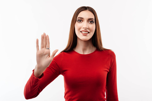 Hello. Portrait of adorable friendly woman with long chestnut hair in casual red sweater waving hand, looking at camera with engaging toothy smile. Studio shot, white background.Human emotions concept - Photo, Image