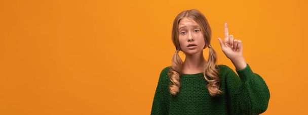 Πορτρέτο της σοβαρής ξανθιά κοπέλα 12-14 ετών σε ζεστό πράσινο πουλόβερ δείχνει δείκτη δάχτυλο, κατσάδα ή λέει μακριά κάποιον. Το κορίτσι σηκώνει το δάχτυλο και λέει: Studio πυροβόλησε κίτρινο φόντο - Φωτογραφία, εικόνα