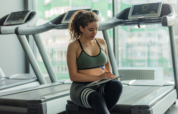 A young fit woman writes information in a training book for further progress in training in gym. Healthy lifestyle concept. Your workout plan - Photo, Image