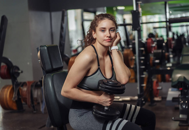 Tired young fit woman holding dumbbells and resting while sitting on a bench in the gym - Фото, зображення