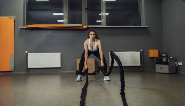 Mujer en forma joven haciendo ejercicio con cuerdas de batalla en clase de entrenamiento. Entrenamiento funcional, proceso de entrenamiento. - Foto, imagen