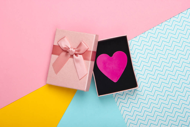 Κουτί δώρου με φιόγκο και διακοσμητική καρδιά σε χρωματιστό φόντο. Ημέρα του Αγίου Βαλεντίνου. Παστέλ χρώμα τάση. Άνω όψη - Φωτογραφία, εικόνα