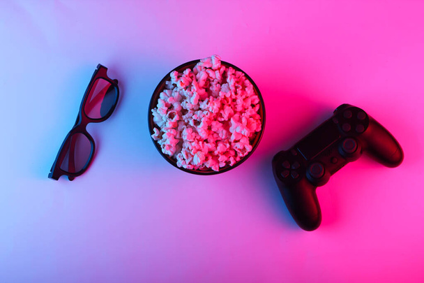 Gamepads, tv remote, 3d glasses and a bowl of popcorn. Neon pink blue light. Gaming, leisure and entertainment concept. Top view - Photo, Image