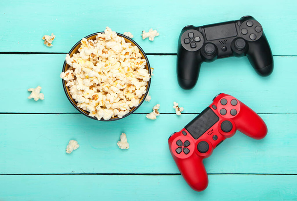 Twee gamepads en een kom popcorn op blauwe houten achtergrond. Gaming, vrije tijd en entertainment concept. Bovenaanzicht - Foto, afbeelding
