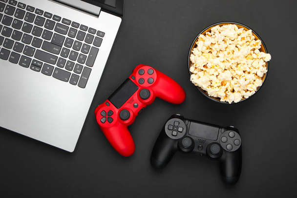 Ноутбук, два геймпада и миска попкорна на черном фоне. Концепция игр, отдыха и развлечений. Вид сверху - Фото, изображение