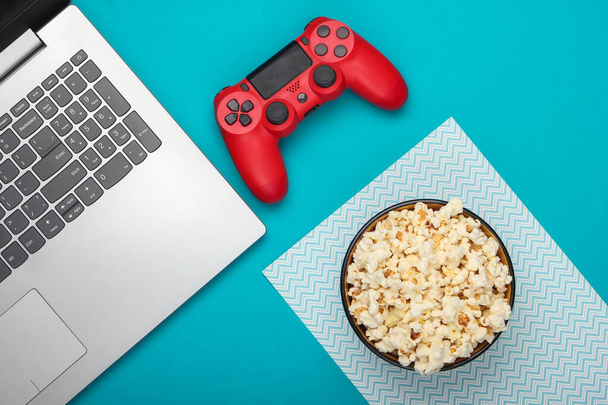Laptop, gamepad en een kom popcorn op blauwe achtergrond. Gaming, vrije tijd en entertainment concept. Bovenaanzicht - Foto, afbeelding