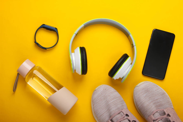 Flat lay composition of modern gadgets for sports and fitness. Wireless headphones, smart bracelet, smartphone, running shoes, water bottle on a yellow background. Top view - Photo, Image