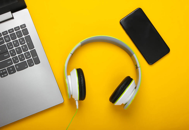 Laptop com fones de ouvido estéreo e smartphone em fundo amarelo. Composição de leigos planos. Aparelhos modernos. Vista superior - Foto, Imagem