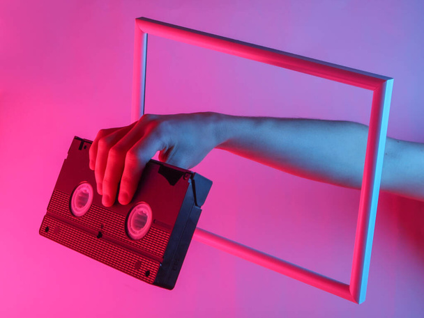 Creative art. Female hand holding video cassette through a soaring frame with neon holographic light. Gradient glow. - Photo, Image