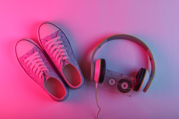 Retro media i rozrywka lat 80-tych. Słuchawki, kasety audio i tenisówki. Neonowe światło gradientowe różowo-niebieskie. Fala wsteczna. Widok z góry - Zdjęcie, obraz