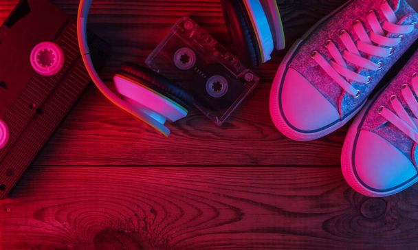 Fones de ouvido estéreo, cassete de áudio e vídeo, tênis em fundo de madeira. Luz azul vermelha néon. Brilho de gradiente. Mídia retrô e entretenimento. Vista superior - Foto, Imagem