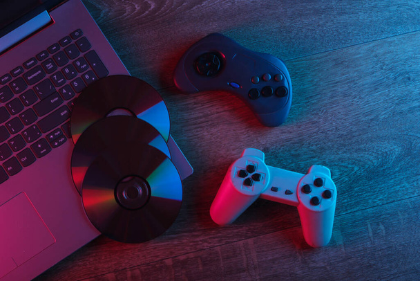 Laptop com gamepads, discos de cd em um piso de madeira. Luz de néon vermelho azul. Brilho de gradiente. Jogos ainda vida. Vista superior - Foto, Imagem