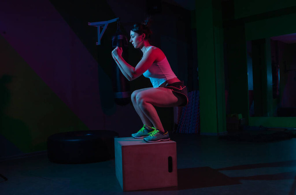 Jonge fit vrouw hurkt op een houten doos in een neon gradiënt roze en blauw licht op een donkere achtergrond. Functioneel opleidingsconcept. - Foto, afbeelding