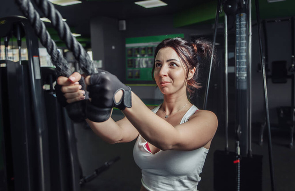 Νεαρή λεπτή γυναίκα εξάσκηση επέκταση των όπλων με σχοινιά στο μηχάνημα άσκησης στο γυμναστήριο - Φωτογραφία, εικόνα