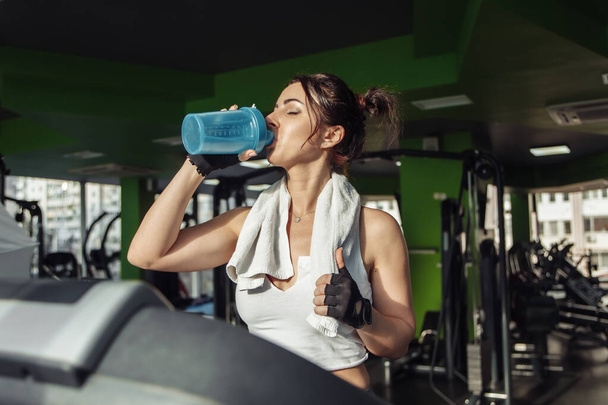 Joven mujer en forma con una toalla en los hombros bebe agua en una cinta de correr. Concepto de pérdida de peso, entrenamiento aeróbico - Foto, imagen