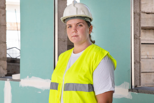 Aantrekkelijke vrouwelijke architect met witte helm en veiligheidsvest die recht naar de camera kijkt tijdens een bezoek aan een bouwplaats en de status en uitvoering van de bouwontwikkeling controleert - Foto, afbeelding