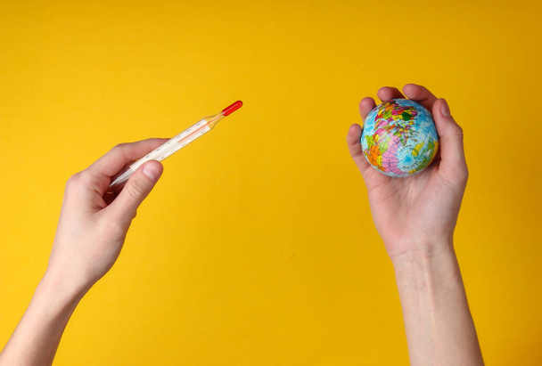 Global warming. Female hands holding a thermometer and a globe on yellow background. Top view - Photo, Image